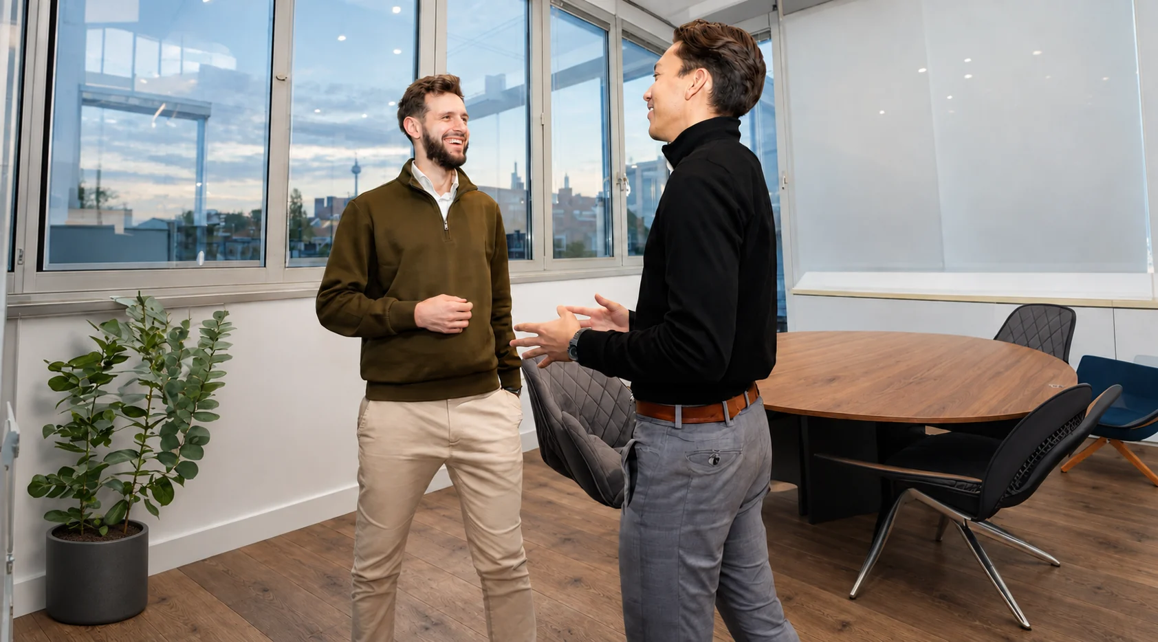 Two colleagues in a bright meeting room with warm oak flooring, a wooden table and dark chairs – digitally enhanced office design